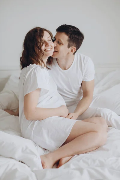 Happy pregnant couple relaxing on white bed and holding belly bu