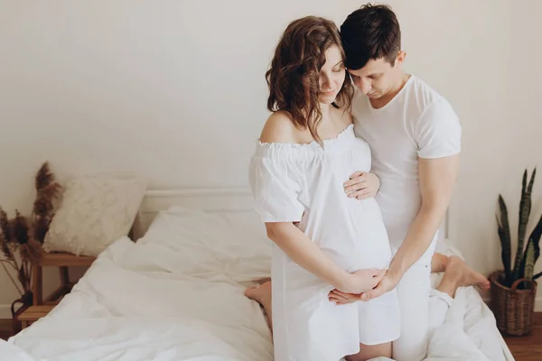 Happy νεαρό ζευγάρι έγκυος κρατώντας την κοιλιά χτύπημα σε λευκό κρεβάτι. STY — Φωτογραφία Αρχείου