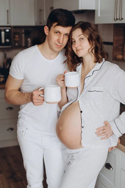 Joyeux jeune couple enceinte en pyjama blanc tenant des tasses et re — Photo