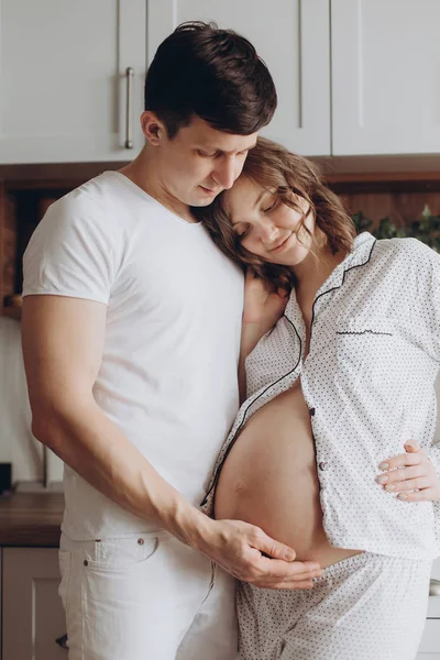 Happy νεαρό ζευγάρι έγκυος στο λευκό πιτζάμες κρατώντας την κοιλιά χτύπημα — Φωτογραφία Αρχείου