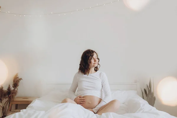 Szczęśliwy w ciąży kobieta w bieli, trzymając wypukłości brzucha i relaksując się na — Zdjęcie stockowe