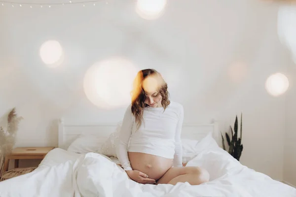 Happy pregnant woman in white holding belly bump and relaxing on — Stock Photo, Image