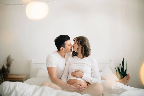 Ευτυχισμένο ζευγάρι έγκυος χαλαρώνετε στο κρεβάτι white και κρατώντας την κοιλιά bu — Φωτογραφία Αρχείου