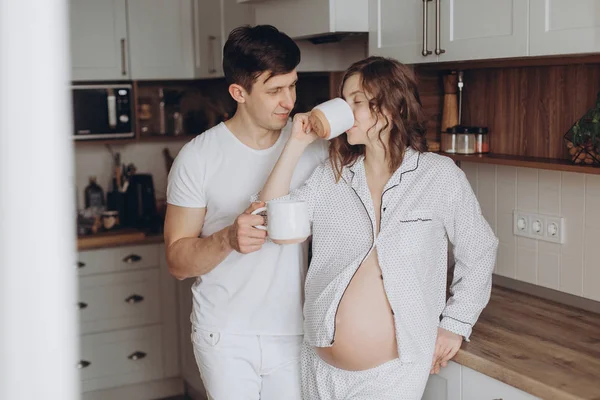 Happy νεαρό ζευγάρι έγκυος στο λευκό πιτζάμες που κατέχουν φλιτζάνια και re — Φωτογραφία Αρχείου