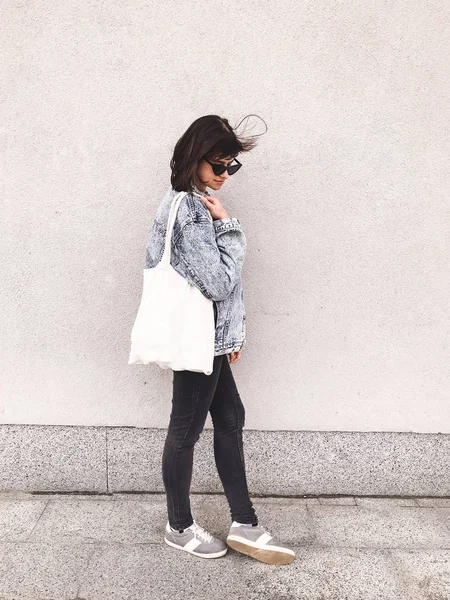 Стильная хипстерша в джинсовой одежде и сумке, позирующая в городе — стоковое фото