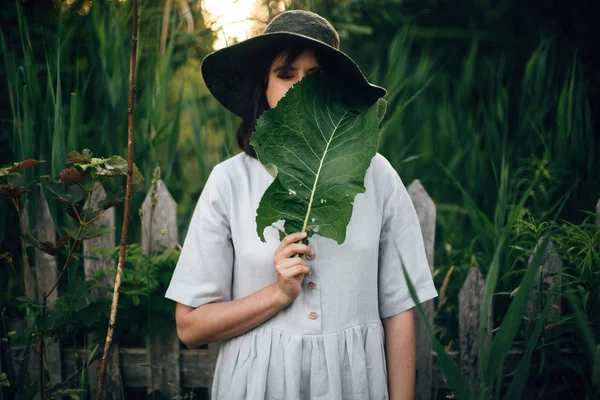 Gadis bergaya dalam gaun linen memegang daun hijau besar di wajah di wo — Stok Foto