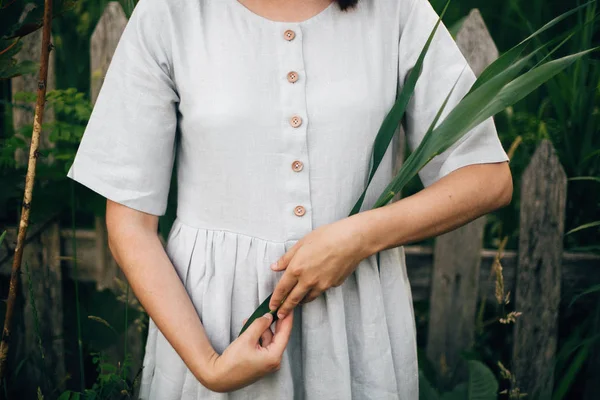 Стильная девушка в льняном платье, держащая зеленый лист у деревянного забора — стоковое фото