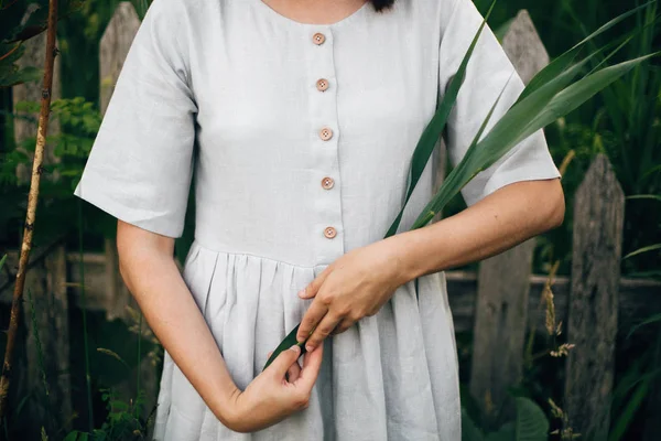 Stylish girl in linen dress holding green leaf at wooden fence a — Stock Photo, Image