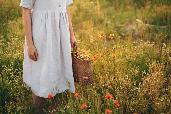 Stylish girl in linen dress holding rustic straw basket with poppy flowers in meadow in sunset light. Boho woman relaxing and gathering wildflowers in summer field. Atmospheric moment