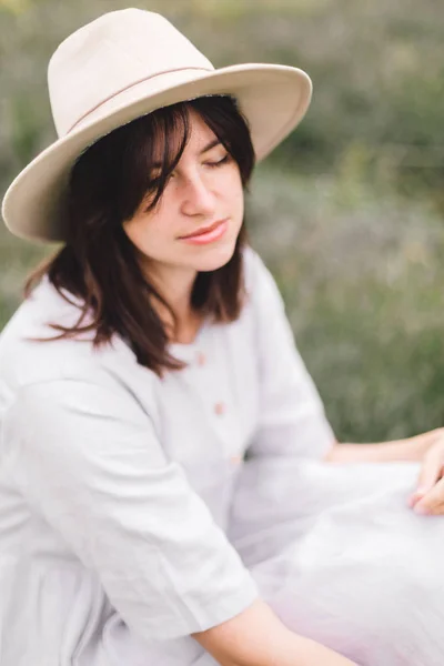 Stylish hipster girl in linen dress and hat sitting on bench at — Stock Photo, Image