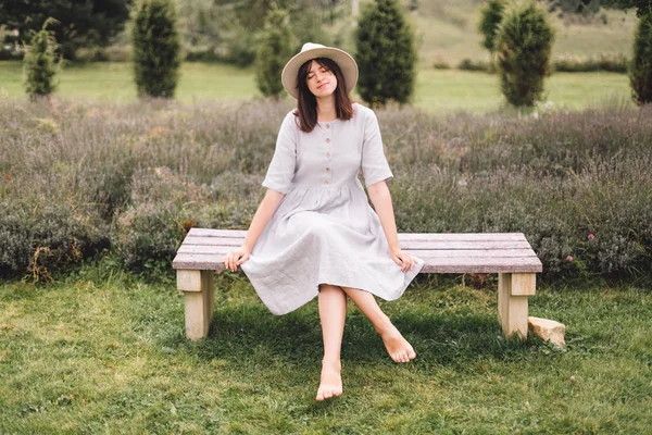 Стильная хипстерша в льняном платье и шляпе сидит на скамейке у — стоковое фото