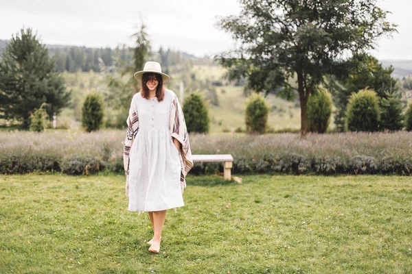 Стильная хипстерша в льняном платье и шляпе ходит по лаванде — стоковое фото