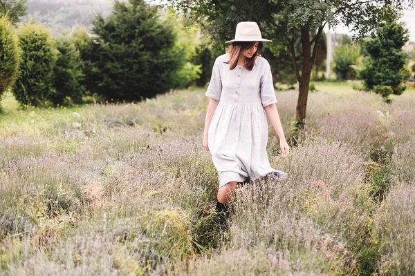 Стильная хипстерша в льняном платье и шляпе ходит в лаванде — стоковое фото