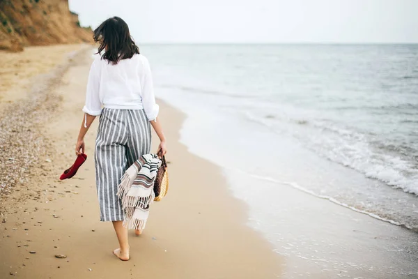 Stylisches Hipstermädchen, das barfuß am Strand läuft, Tasche in der Hand hält und — Stockfoto