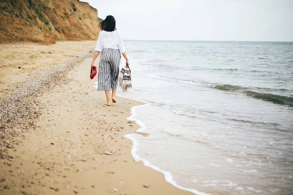 Стильная хипстерша ходит босиком по пляжу, держа сумку и — стоковое фото