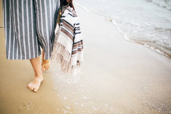 Стильная девушка-хипстер, идущая босиком по пляжу, держа в руках сумку — стоковое фото