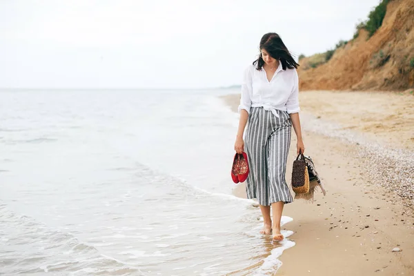 Stílusos csípő lány sétáló mezítláb a strandon, gazdaság táska és — Stock Fotó