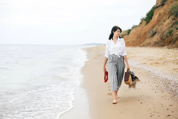 Стильная хипстерша ходит босиком по пляжу, держа сумку и — стоковое фото