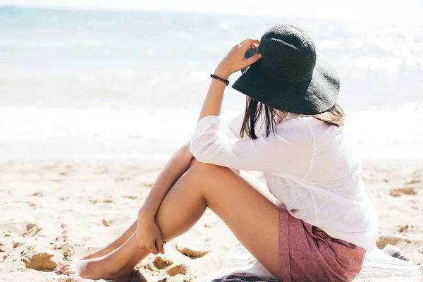 Stylisches Hipster-Mädchen mit Hut sitzt am Strand und bräunt sich in der Nähe — Stockfoto