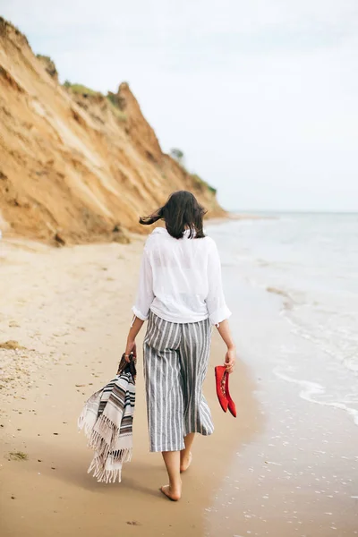 Stylisches Hipstermädchen, das barfuß am Strand läuft, Tasche in der Hand hält und — Stockfoto