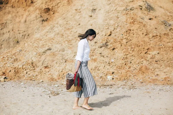 Stylish hipster girl walking barefoot on beach, holding bag and — Stock Photo, Image