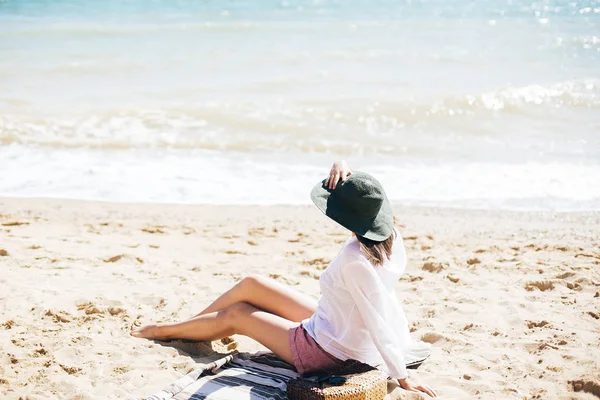 Menina elegante hipster em chapéu sentado na praia e bronzeamento perto de se — Fotografia de Stock