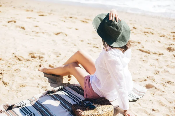 Stylisches Hipster-Mädchen mit Hut am Strand sitzend mit Strohtasche und — Stockfoto