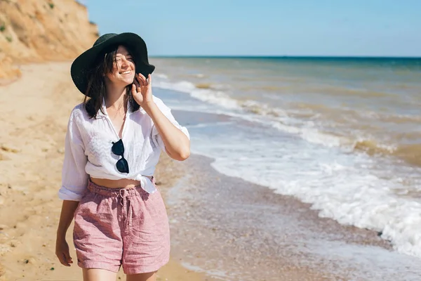 Stylisches Hipster-Mädchen mit Hut, das am Strand spazieren geht und lächelt. Sommer — Stockfoto