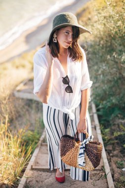 Şık boho kız s çim ile kumlu uçurumda ayakta şapka