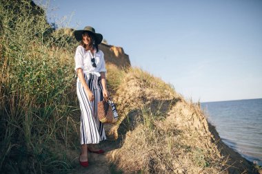 Şık boho kız s çim ile kumlu uçurumda ayakta şapka