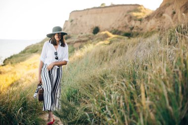 Şapka şık boho kız yakın çim ile kumlu uçurumda yürüyüş 