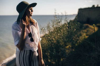 Şık boho kız s çim ile kumlu uçurumda ayakta şapka