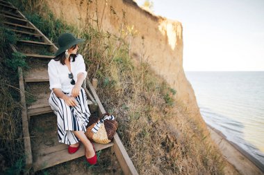 Güneşli ışıkta ahşap merdivenlerde oturan şapkaşık boho kız