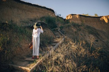 Şapka şık boho kız deniz kenarında kumlu uçurumun fotoğrafını çekiyor