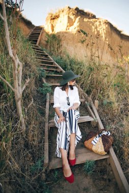 Güneşli ışıkta ahşap merdivenlerde oturan şapkaşık boho kız