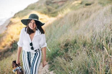 Şapka şık boho kız yakın çim ile kumlu uçurumda yürüyüş 