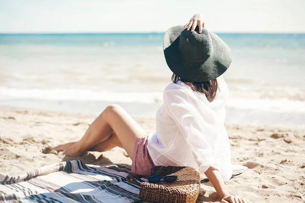 Stylisches Hipster-Mädchen mit Hut am Strand sitzend mit Strohtasche und — Stockfoto