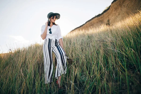 Stylish boho girl in hat posing at sandy cliff with grass near s — Stock Photo, Image