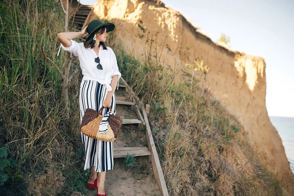 Stylisches Boho-Mädchen mit Hut posiert an sandiger Klippe mit Gras in der Nähe s — Stockfoto