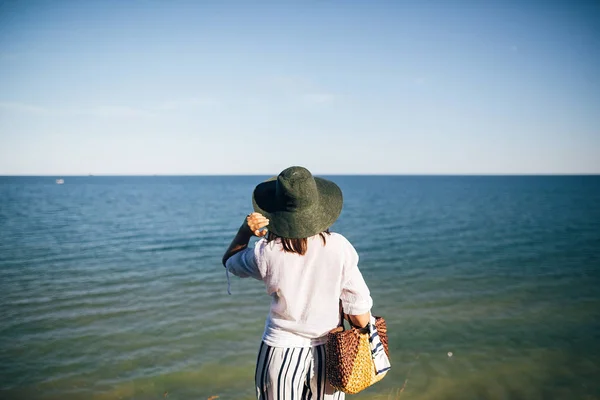 Стильная девушка в шляпе, глядящая на море в солнечном вечернем свете. — стоковое фото