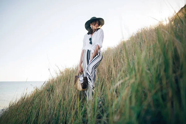 Stylish boho girl in hat posing among grass in sunny evening lig — Stock Photo, Image