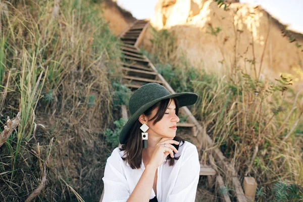 Стильная девушка в шляпе, сидящая на деревянной лестнице в солнечном свете — стоковое фото