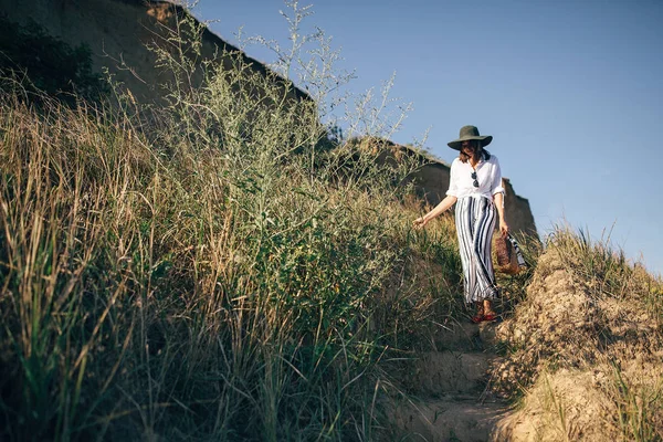 Stylish boho girl in hat walking at sandy cliff with grass near — Stock Photo, Image