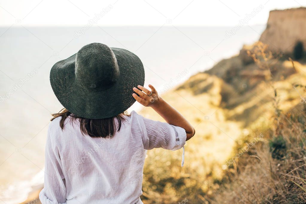 Stylish boho girl in hat looking at sea in sunny evening light f