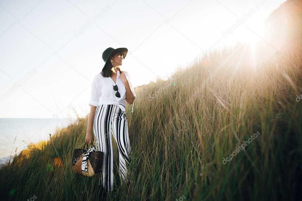 Stylish boho girl in hat posing among grass in sunny evening lig