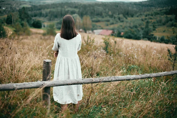 Κομψό κορίτσι σε λινό φόρεμα στέκεται σε παλαιωμένο ξύλινο φράχτη μεταξύ — Φωτογραφία Αρχείου
