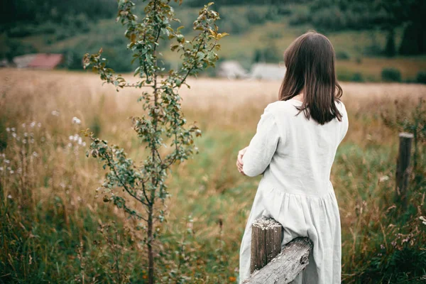 Стильная девушка в льняном платье стоит у стареющего деревянного забора среди — стоковое фото