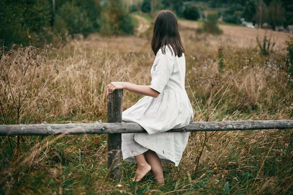 Stylish girl in linen dress sitting on aged wooden fence among h — Stock Photo, Image