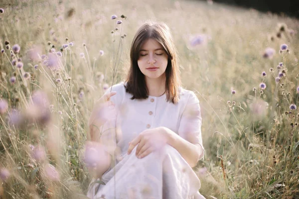 Stijlvolle meisje in rustieke jurk en hoed zittend onder wilde bloemen a — Stockfoto