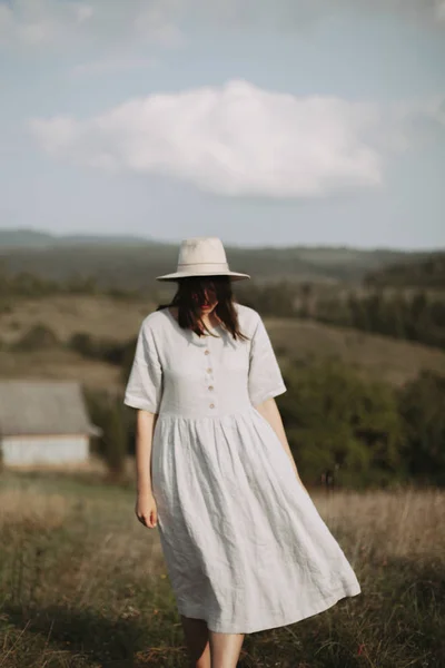 Stylish girl in linen dress and hat walking barefoot in grass in — Stock Photo, Image